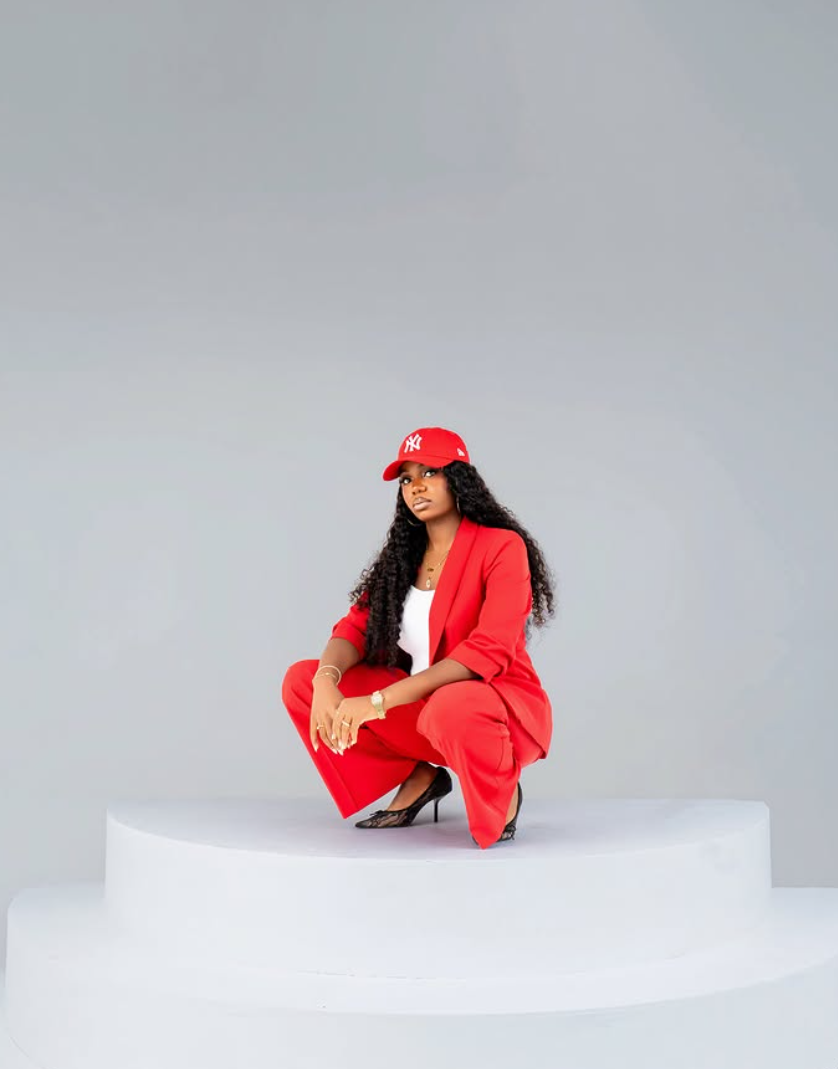 Studio portrait of a woman in a red tailored suit and cap crouching on a white plinth